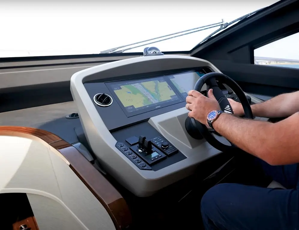 A person with a watch steers a yacht, with a modern dashboard displaying navigation maps and control buttons.